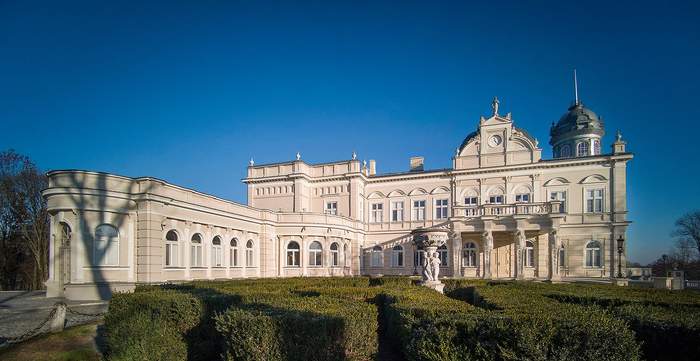 Pokaż zdjęcie: Duży dwukondygnacyjny obiekt z wieżą zegarową oraz kopułą po prawej stronie. Lewa część budynku, która jest prostopadła do frontu pałacu, posiada tylko jedną kondygnację. W tle widoczne jest bezchmurne niebo. Przed pałacem można dostrzec żywopłot z bukszpanu.